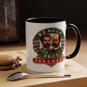 Puede incluir: Una taza de cerámica blanca con interior y asa negros. La taza presenta un logotipo circular con retratos de dos hombres, las palabras "BEAGLE BROS SOFTWARE" y el año "1984". La taza está sobre una superficie de madera con una cuchara y un pastel.