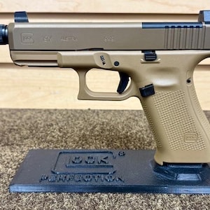 Puede incluir: Una pistola Glock 19X de color arena con un silenciador negro y miras negras. La pistola está en un soporte negro con las palabras "GLOCK Perfection" impresas en él.