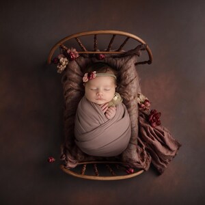 May include: A newborn baby girl sleeping in a wooden crib with a floral patterned blanket. The baby is wearing a light brown swaddle and a flower headband.