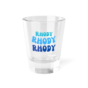 May include: A clear glass shot glass with the word "RHODY" printed three times in blue, teal, and light blue.