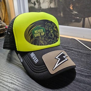 May include: A neon yellow and black trucker hat featuring a black mesh back and a front panel with a graphic of a warrior and the text NEVER GIVE UP. The black brim displays the word NEVER and a tan patch with a white lightning bolt icon.