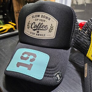 May include: A black trucker hat featuring a beige patch with the text SLOW DOWN SIP YOUR Coffee STAY AWHILE and a light blue patch on the brim displaying the number 19 in dark gray. A small black label reads STRONG COFFEE STRONG MIND.