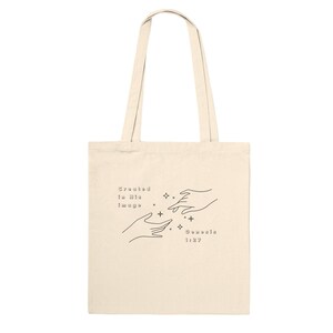 May include: Cream-colored tote bag with black line art of two hands reaching towards each other, with stars and the text "Created in His Image, Genesis 1:27". The bag has long handles.