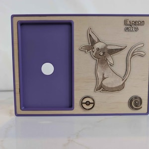 May include: A wooden card holder with a purple frame and a light wood panel. The panel features a laser-cut illustration of an Espeon character, the text "Espeon (#0196)", and two circular designs. A white circle is in the purple frame.