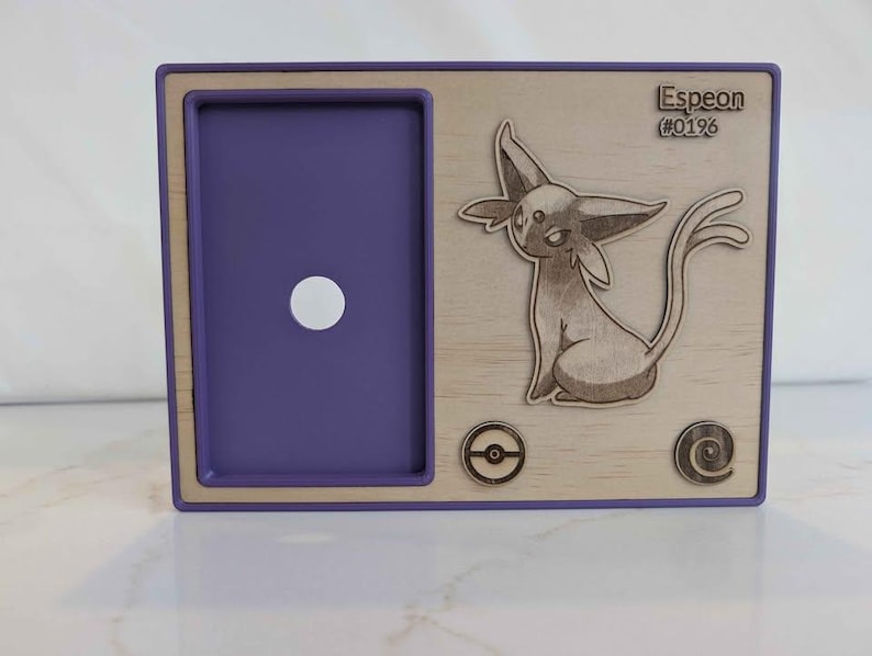 May include: A wooden card holder with a purple frame and a light wood panel. The panel features a laser-cut illustration of an Espeon character, the text "Espeon (#0196)", and two circular designs. A white circle is in the purple frame.