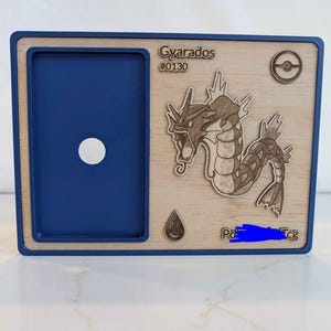 May include: A blue and wood-effect card holder with a laser-cut image of Gyarados, a Pok&eacute;mon character. The holder has a blue frame and a circular opening. The text "Gyarados #0130" is visible.