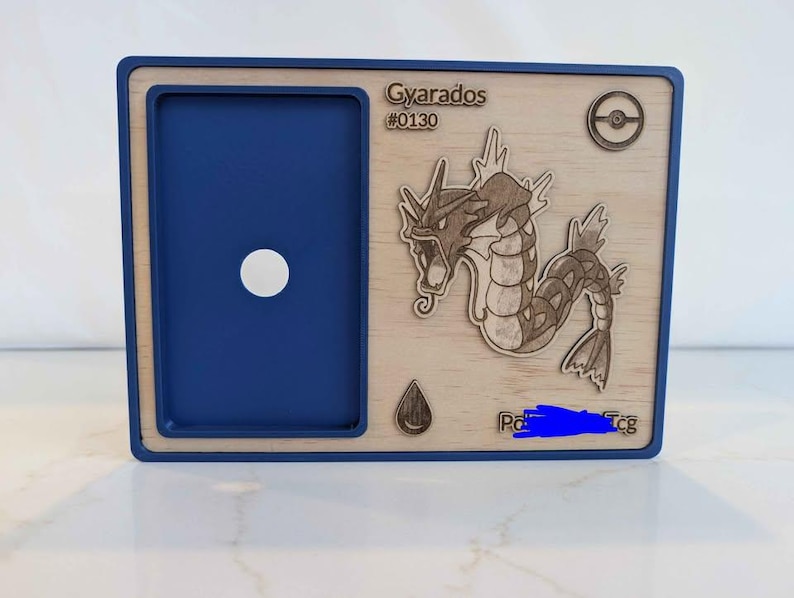May include: A blue and wood-effect card holder with a laser-cut image of Gyarados, a Pok&eacute;mon character. The holder has a blue frame and a circular opening. The text "Gyarados #0130" is visible.