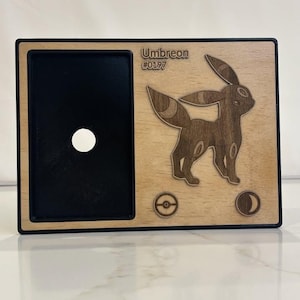 May include: Wooden Umbreon Pok&eacute;mon card holder with a black interior and a light wood exterior. The design features the Umbreon character, the name "Umbreon" and the number "#0197". Includes a Pok&eacute; Ball and moon symbol.