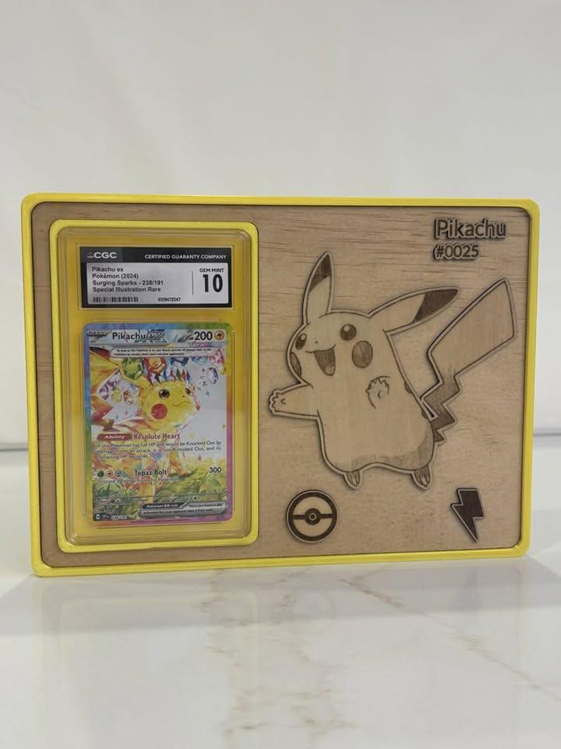 May include: A wooden display frame with a yellow border, holding a graded Pikachu Pok&eacute;mon card. The card is in a protective case, graded as Gem Mint 10. A Pikachu illustration and text "Pikachu #0025" are etched on the wood.