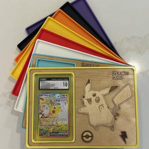 May include: A wooden display featuring a Pikachu design and a graded Pok&eacute;mon card. The display has a yellow frame and includes the text "Pikachu #0025". Various coloured trays are stacked behind the display.