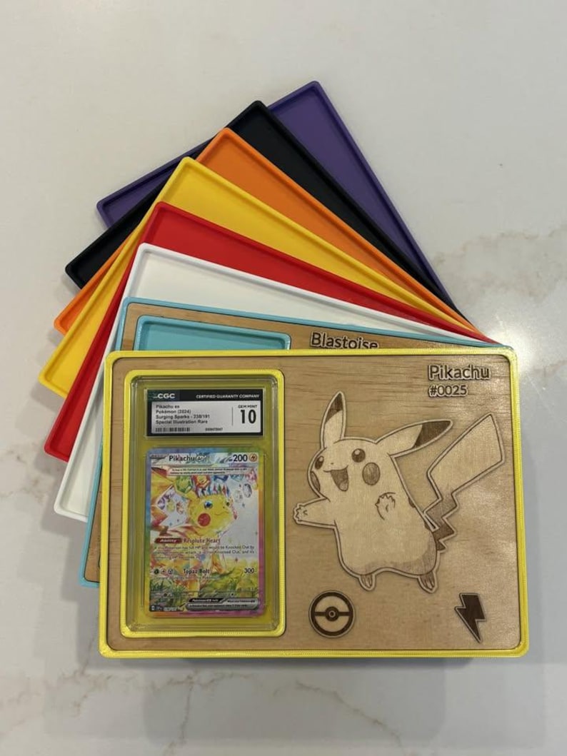 May include: A wooden display featuring a Pikachu design and a graded Pok&eacute;mon card. The display has a yellow frame and includes the text "Pikachu #0025". Various coloured trays are stacked behind the display.