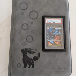 May include: A gray trading card binder featuring an embossed Umbreon character and circular patterns on the cover. A black frame holds a PSA-graded 2025 Pokemon Mega Charizard X ex trading card. The bottom right corner displays the TCGUARD logo.