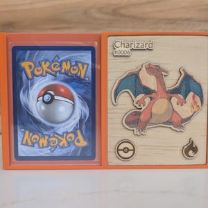 May include: An orange display frame holding a Pokémon card on the left and a wooden plaque on the right. The plaque features a laser-cut Charizard illustration, the text Charizard #0006, a Poké Ball icon, and a fire symbol icon on a light wood grain.