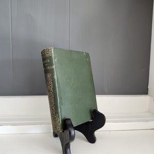 Könnte beinhalten: Ein Vintage-Buch mit dem Titel "Days of the Year" in einem dunkelgrünen Einband, präsentiert auf einem schwarzen Holzständer. Der Buchrücken weist goldene dekorative Elemente auf. Der Hintergrund ist eine neutrale graue Wand und weiße Zierleisten.