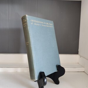 May include: A hardcover book titled A Little Book of Western Verse by Eugene Field with a muted blue-green cover and gold lettering, displayed upright on a black wooden stand. The title and author name are printed on the front and spine.