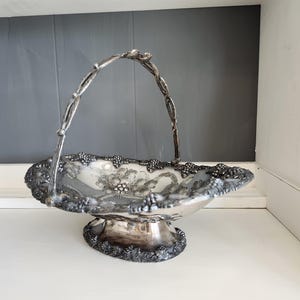 Könnte beinhalten: Ein silberfarbener, verzierter Obstkorb mit Henkel. Der Korb hat einen gewellten Rand mit Trauben-Details. Der Innenraum hat ein Blumendesign. Der Sockel ist ebenfalls mit Traubenblatt- und Traubenmotiven verziert.