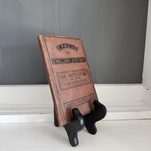 Puede incluir: Un libro antiguo titulado "Epochs of English History" sobre un soporte de exhibición de madera negra. El libro tiene una cubierta marrón con texto negro. El soporte está sobre una superficie blanca con un fondo gris.