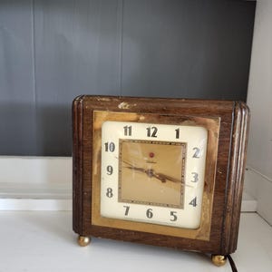 以下が含まれることがあります： 金色文字盤と黒色の針を持つ、木製のビンテージ時計。時計の12時位置には赤い点があり、文字盤には「Telechron」と印刷されています。