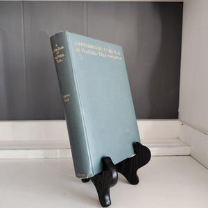 May include: A light blue hardcover book titled A Little Book of Profitable Tales by Eugene Field, resting on a black display stand. The front cover and spine feature gold lettering. The book is positioned upright to show the cover and spine.