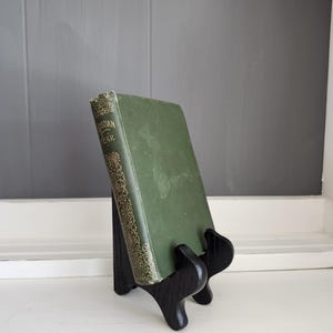 May include: A vintage green book titled "Christian Year" is displayed on a black wooden stand. The book has gold lettering and decorative accents. The stand is positioned on a white surface against a gray background.