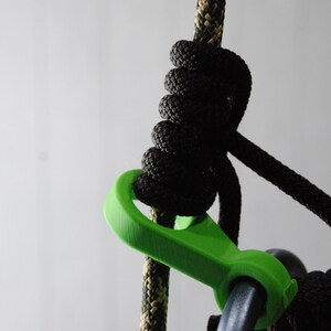 May include: Close-up of a climbing rope with a black knot and a bright green carabiner. The rope is black and tan, and the carabiner is attached to a metal ring. The background is a plain, light gray.
