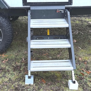 May include: A three-step, black metal RV step with a gray, non-slip tread. The step is labeled "Lippert" and "Slide Step". The step is extended and resting on two white concrete blocks.