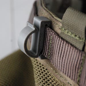 Może przedstawiać: Zbliżenie szarego plastikowego klipsa przymocowanego do brązowego i oliwkowozielonego paska materiału. Klips ma zakrzywiony uchwyt i prostokątną podstawę. Materiał ma tkaną fakturę.