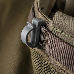 May include: A grey plastic C-shaped clip attached to a brown nylon webbing strap. The strap is integrated into a brown mesh fabric panel. The clip features a curved hook design and is secured firmly onto the horizontal textile webbing loop.