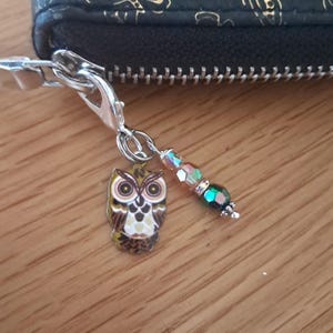 May include: A silver-toned metal zip pull charm attached to a black wallet. The charm features a small, flat owl pendant with brown, cream, and yellow enamel details, accompanied by a short strand of three faceted, iridescent glass beads.