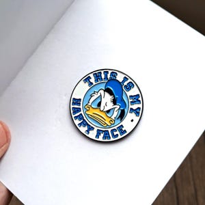 May include: A circular enamel pin with a cartoon image of Donald Duck and the text "THIS IS MY HAPPY FACE." The pin has a white border, a light blue background, and a blue hat.