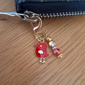 May include: A gold-toned metal lobster clasp zip pull charm attached to a dark wallet. The charm features a red enamel bird with white wing detail and a dangling beaded accent consisting of clear, gold, and red faceted glass beads.