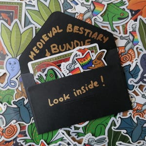Könnte beinhalten: Ein schwarzer Umschlag mit den goldenen Aufschriften "MEDIEVAL BESTIARY BUNDLE" und "Look inside!". Er enthält bunte Aufkleber mit Kreaturen und dem Text "GAY AGENDA". Die Aufkleber sind um den Umschlag herum verstreut.