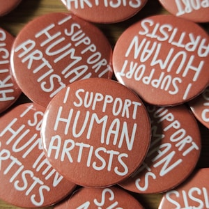 Może przedstawiać: Kolekcja okrągłych przypinek z tłem w kolorze terakoty i białym napisem I SUPPORT HUMAN ARTISTS. Przypinki są ułożone w grupę, a każda z nich zawiera ten sam pogrubiony, bezszeryfowy napis w układzie warstwowym.