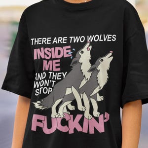 May include: Black t-shirt with a graphic of two howling wolves and the text "THERE ARE TWO WOLVES INSIDE ME AND THEY WON'T STOP FUCKIN'". The wolves are gray and white, with pink text.