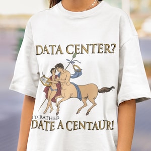 May include: A white short-sleeved t-shirt featuring a graphic illustration of a centaur and a person with a lyre. The text reads DATA CENTER? I'D RATHER DATE A CENTAUR! The design includes muted tones of brown, blue, and red on the fabric.