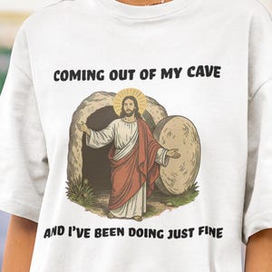 May include: White t-shirt with the text "COMING OUT OF MY CAVE AND I'VE BEEN DOING JUST FINE." The shirt features a graphic of a figure emerging from a cave. The figure is wearing a red and white robe.