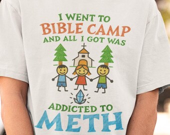 May include: White t-shirt with the text "I went to Bible Camp and all I got was addicted to meth." The shirt features a graphic of stick figures, trees, and a church. The shirt is a light color and has short sleeves.