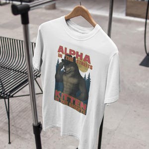 May include: White t-shirt with the text "ALPHA IN THE STREETS, KITTEN IN THE SHEETS" above a cartoon illustration of a muscular wolf. The wolf is flexing its bicep with a forest and moon in the background.