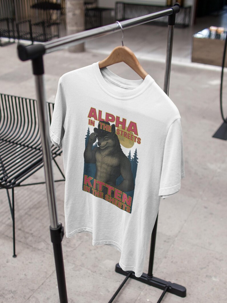 May include: White t-shirt with the text "ALPHA IN THE STREETS, KITTEN IN THE SHEETS" above a cartoon illustration of a muscular wolf. The wolf is flexing its bicep with a forest and moon in the background.