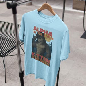 May include: Light blue t-shirt with the text "ALPHA IN THE STREETS, KITTEN IN THE SHEETS" above a cartoon wolf illustration. The wolf is flexing its bicep. The shirt is hanging on a wooden hanger.