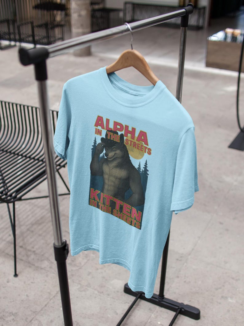 May include: Light blue t-shirt with the text "ALPHA IN THE STREETS, KITTEN IN THE SHEETS" above a cartoon wolf illustration. The wolf is flexing its bicep. The shirt is hanging on a wooden hanger.