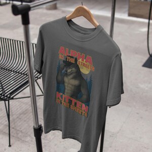 May include: Dark gray t-shirt with the text "ALPHA IN THE STREETS, KITTEN IN THE SHEETS" above and below an illustration of a muscular wolf-like character. The design features a retro style with a forest and moon background.