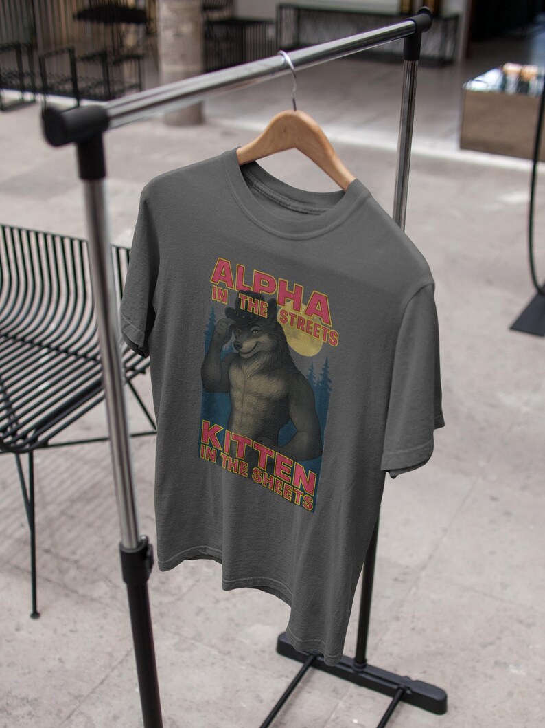 May include: Dark gray t-shirt with the text "ALPHA IN THE STREETS, KITTEN IN THE SHEETS" above and below an illustration of a muscular wolf-like character. The design features a retro style with a forest and moon background.