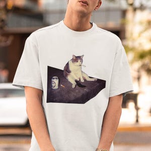 May include: White t-shirt featuring a graphic of a cat with a cigarette, sitting next to a can and a cigarette. The cat is depicted in shades of gray and white. The shirt is a casual, short-sleeved style.