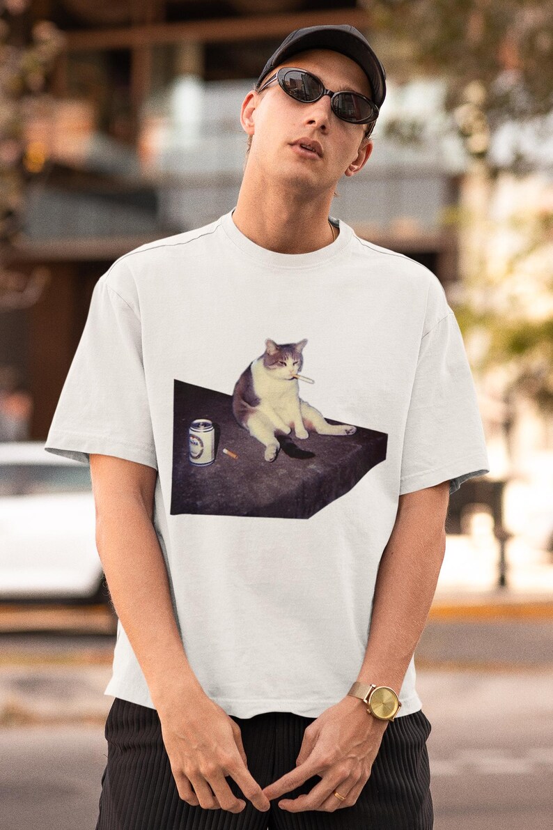 May include: White t-shirt featuring a graphic of a cat with a cigarette, sitting next to a can and a cigarette. The cat is depicted in shades of gray and white. The shirt is a casual, short-sleeved style.