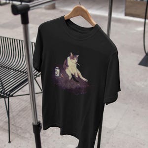 May include: Black t-shirt featuring a meme of a cat sitting with a cigarette and a can. The cat is white and gray. The can is silver with a white label. The shirt is hanging on a wooden hanger on a metal rack.