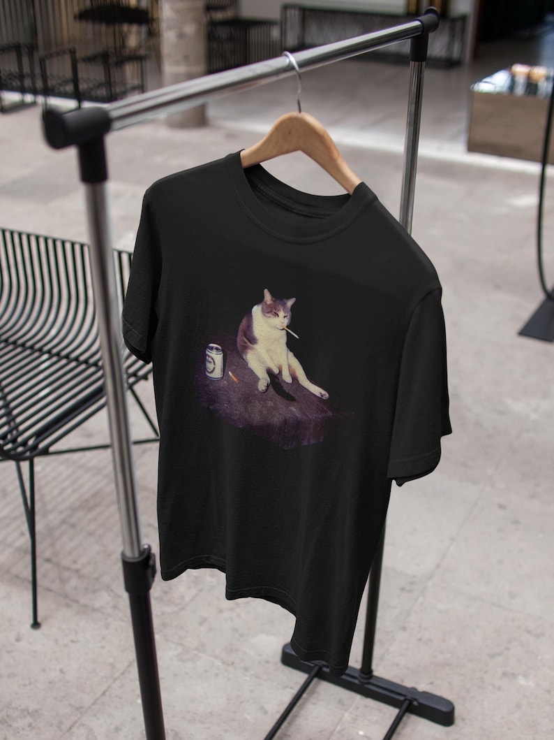 May include: Black t-shirt featuring a meme of a cat sitting with a cigarette and a can. The cat is white and gray. The can is silver with a white label. The shirt is hanging on a wooden hanger on a metal rack.