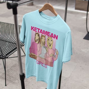 May include: Light blue t-shirt with a graphic of three women with horse heads wearing pink shirts and holding cigarettes. The text "KETAMEAN GIRLS" is printed on the shirt.