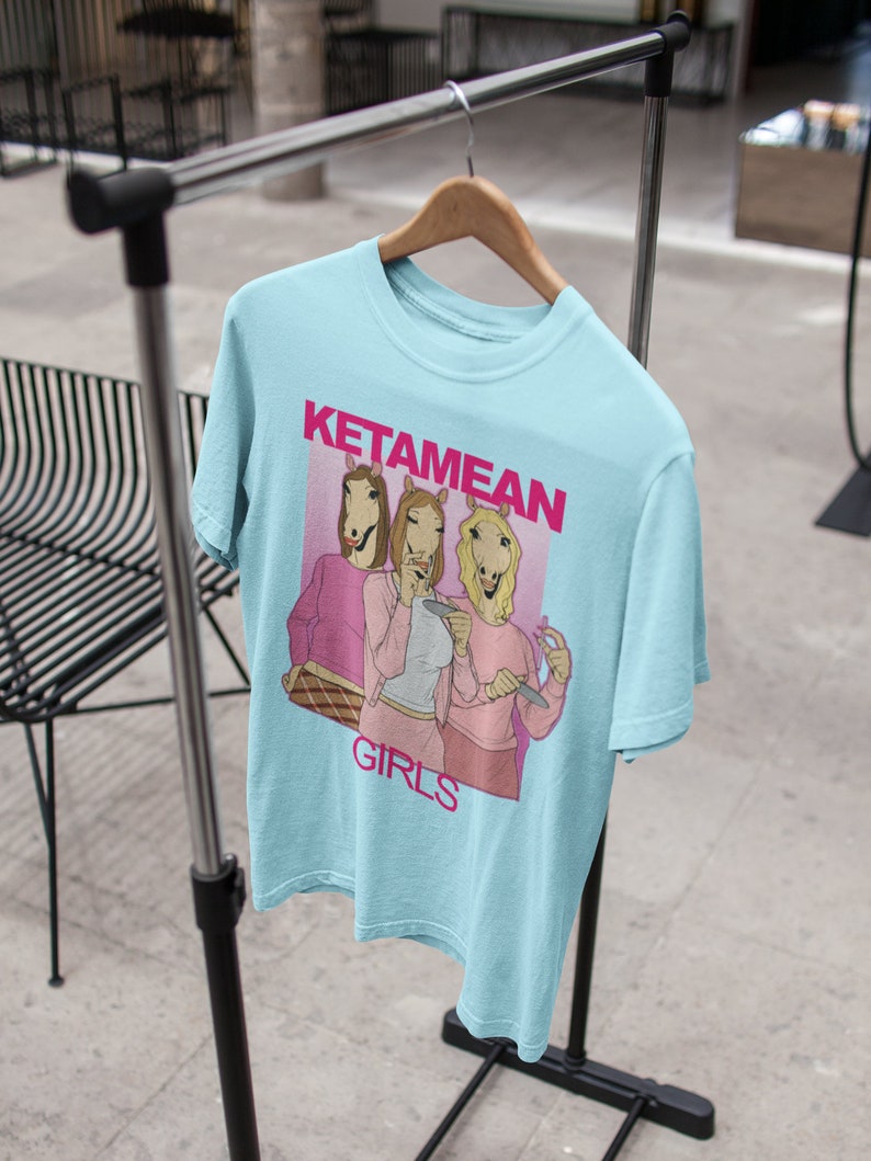 May include: Light blue t-shirt with a graphic of three women with horse heads wearing pink shirts and holding cigarettes. The text "KETAMEAN GIRLS" is printed on the shirt.