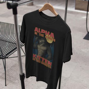 May include: Black t-shirt with a graphic of a muscular wolf-like character under a full moon. Text on the shirt reads "ALPHA IN THE STREETS, KITTEN IN THE SHEETS" in red and yellow lettering. The shirt is hanging on a wooden hanger.